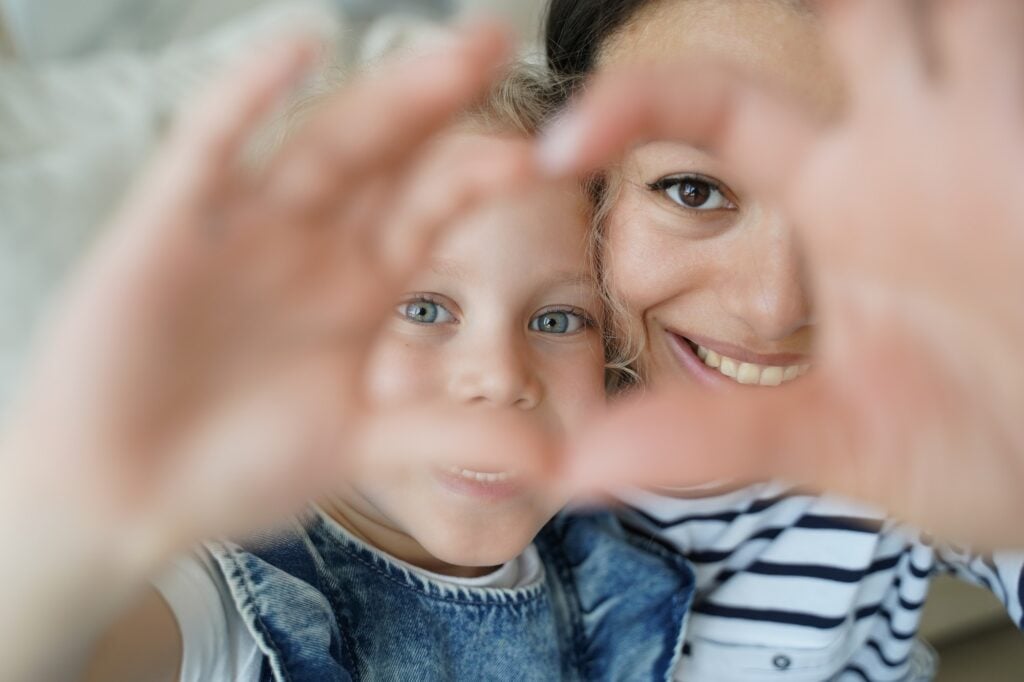 Smiling foster mom, adopted child girl daughter taking selfie, showing love heart gesture together